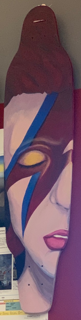 Acrylic painting on a skate deck of a Bowie-inspired face with a blue lightning bolt and a gold-rimmed eye